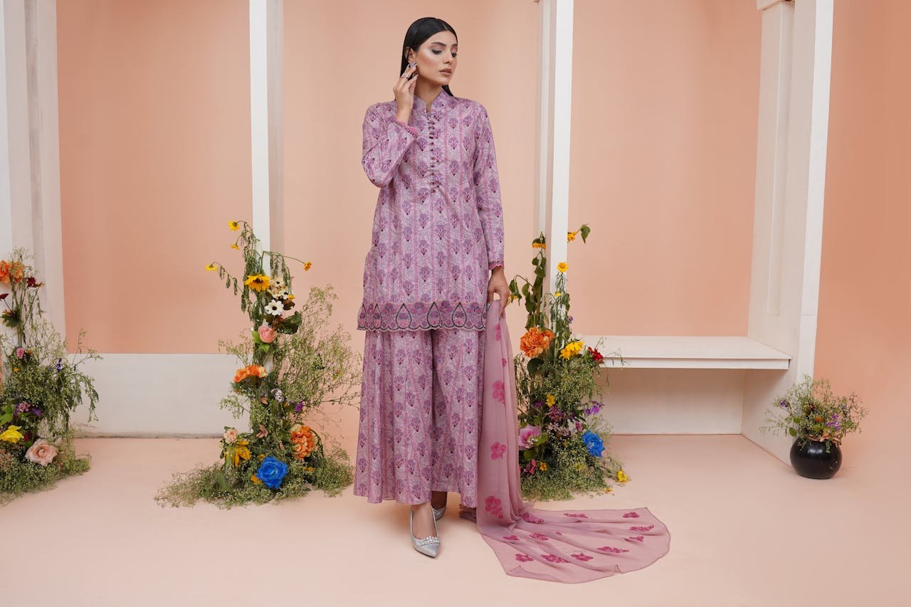 A woman in a purple traditional dress stands elegantly surrounded by vibrant floral decorations.